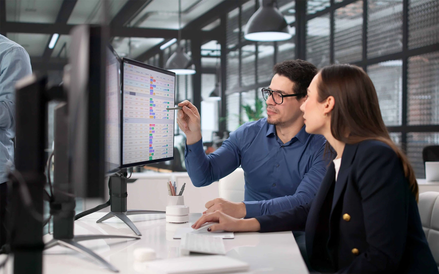 Two professionals sit at a desk in a modern office, reviewing financial or operational data displayed on a computer monitor.