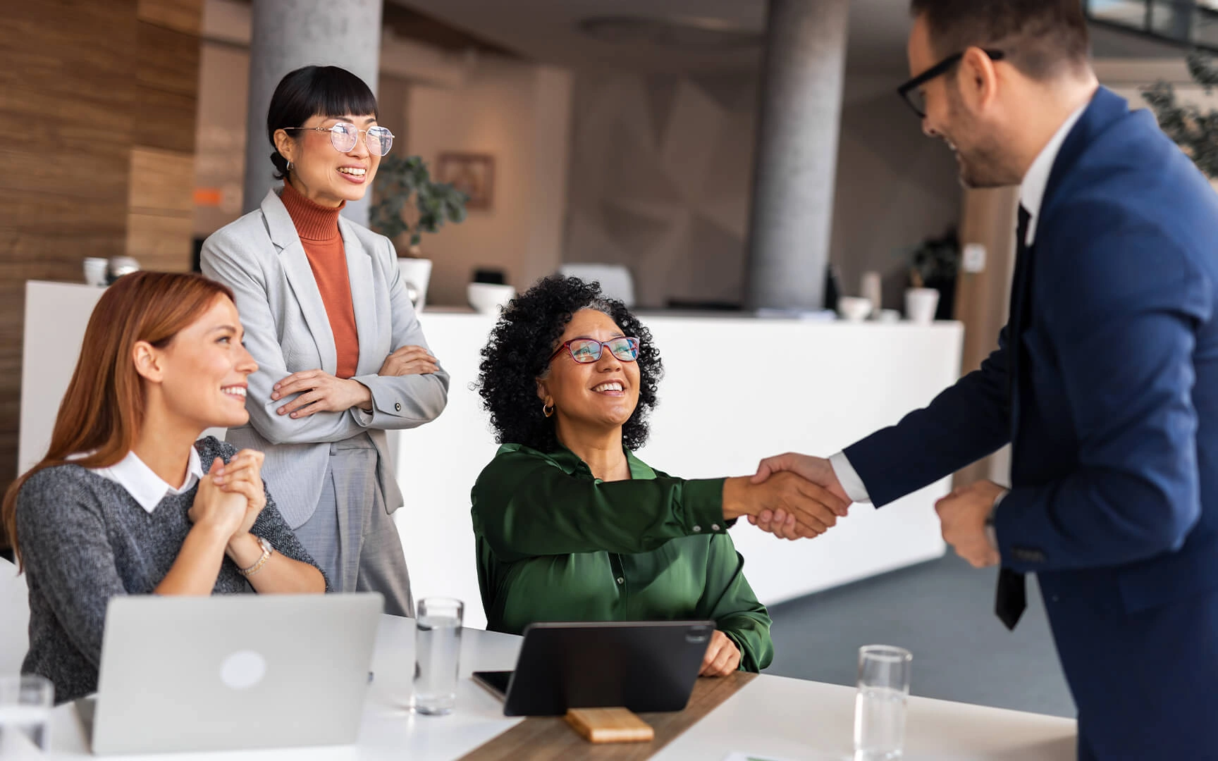 Des professionnels collaborent dans un bureau moderne ; deux personnes se serrent la main sous le regard de leurs collègues, avec des ordinateurs et tablettes à disposition.
