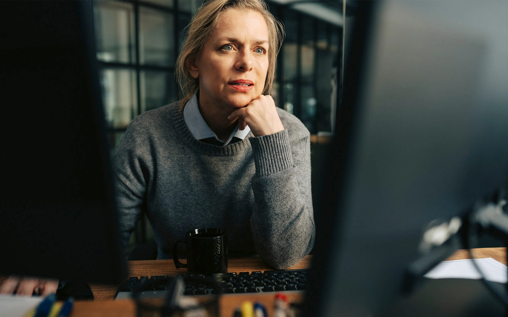 Person seated at a desk looking at a computer screen, with a keyboard, coffee mug, and office items in the foreground.