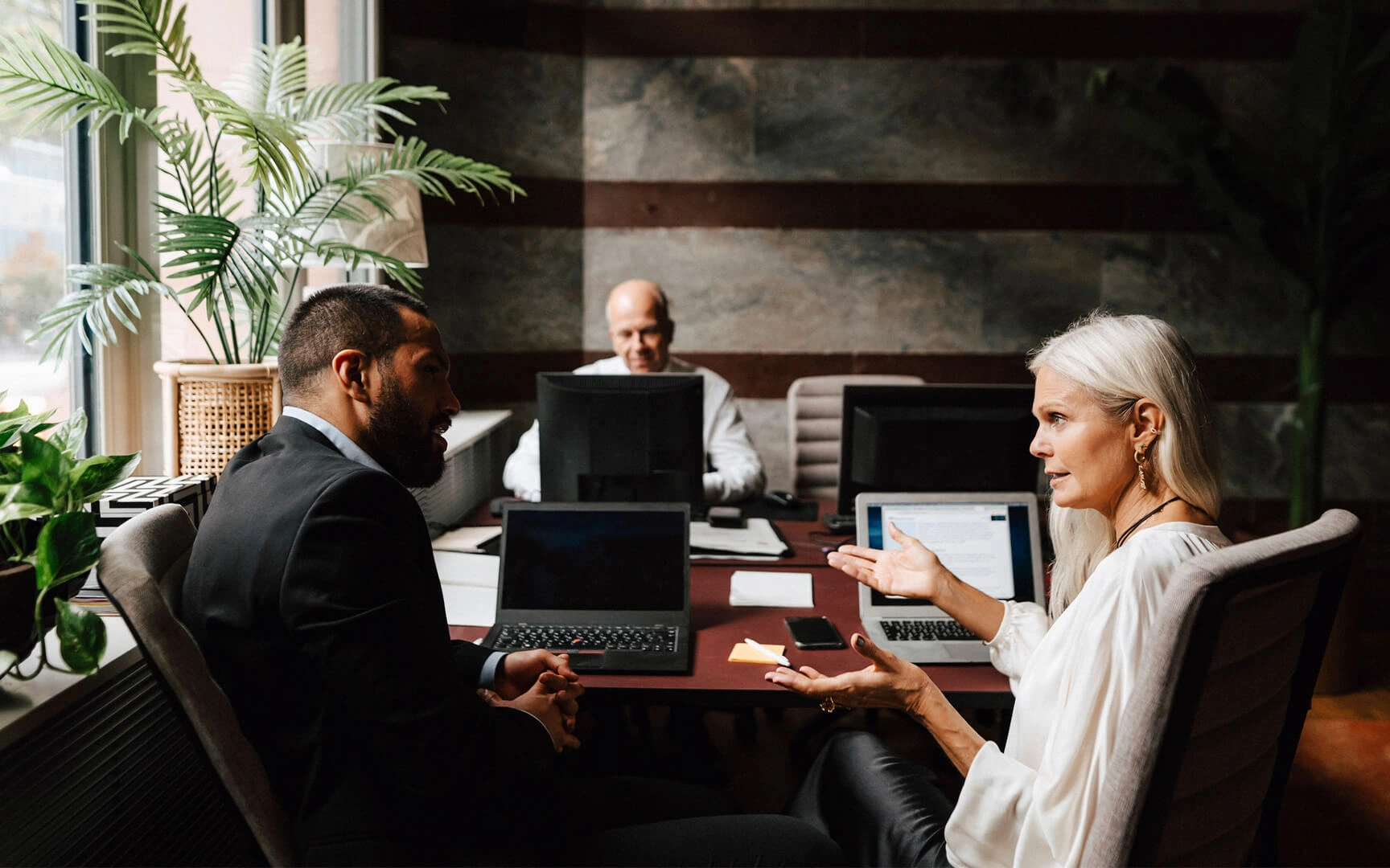 Trois professionnels discutent autour d'une table de réunion avec des ordinateurs portables dans un bureau moderne.