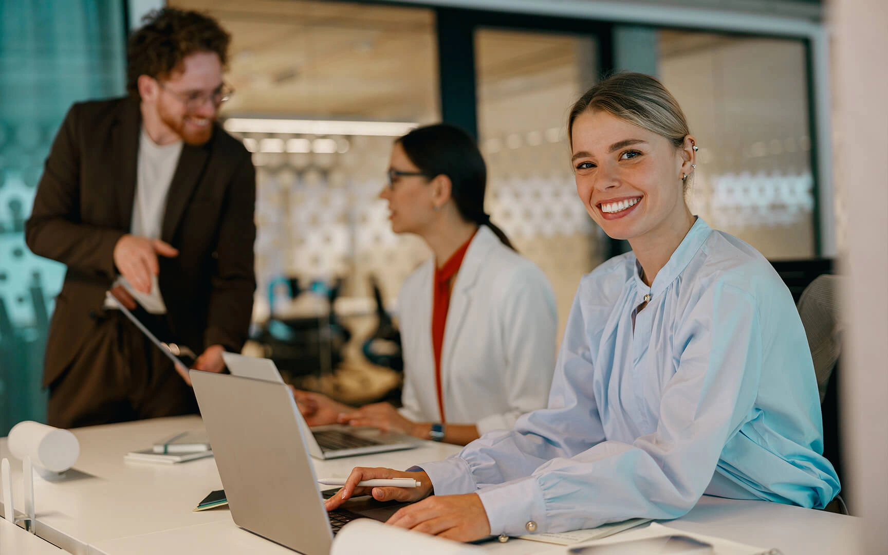 Drei Personen arbeiten an Laptops in einem modernen Büro; eine Person sitzt im Vordergrund an einem Schreibtisch, während zwei Kollegen im Hintergrund zusammenarbeiten.