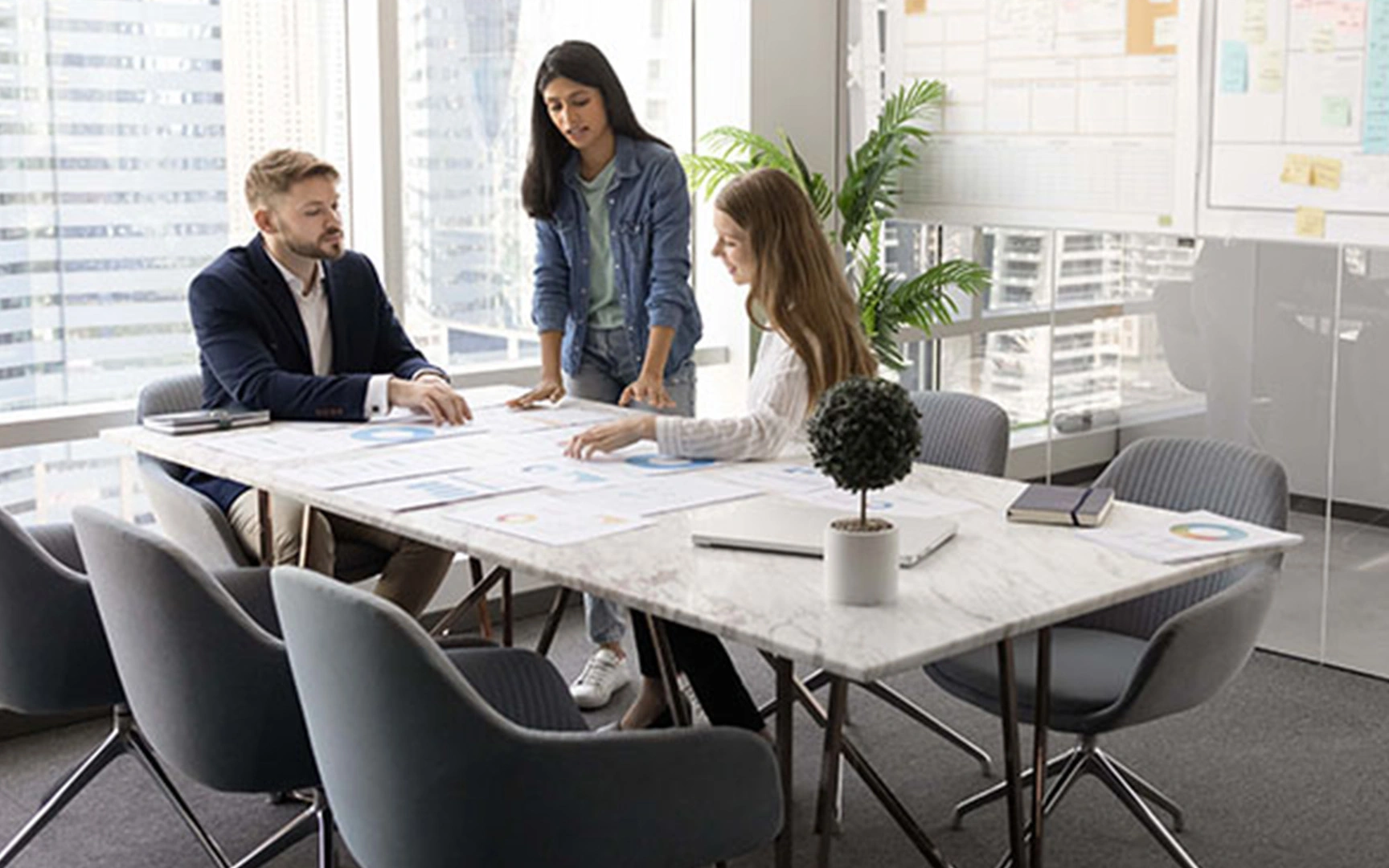 Three professionals collaborating around a table in a modern office, reviewing printed charts and financial data during a planning discussion.