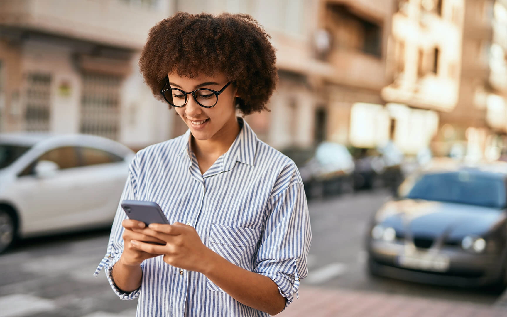 Person standing on a city street, wearing a striped button-down shirt and holding a smartphone, with parked cars and residential buildings in the background.