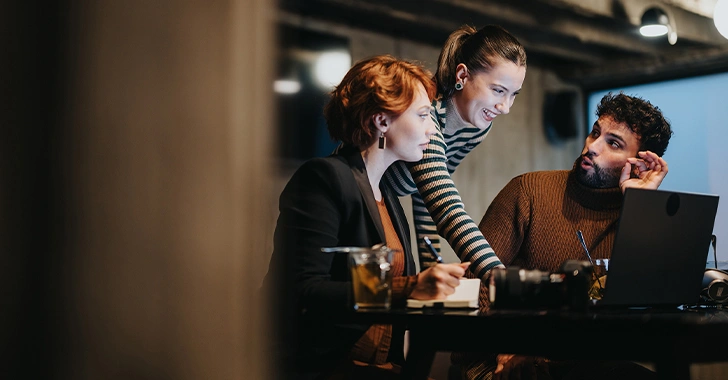 Three people are collaborating at a table with a laptop. There are notebooks and a glass on the table. The group is engaged in conversation in a dimly lit room.