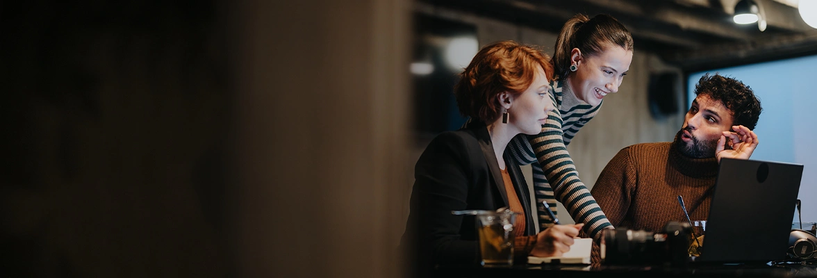 Three people are collaborating at a table with a laptop. There are notebooks and a glass on the table. The group is engaged in conversation in a dimly lit room.