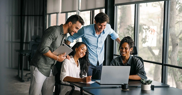 A group of four people in a modern office setting gather around a laptop, engaged and smiling. One person holds a tablet. The room has large windows and a potted plant on the table.