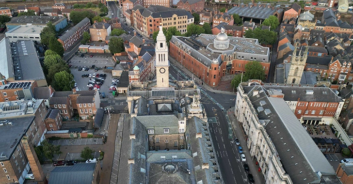 Aerial view of a town with prominent buildings, including a church with a clock tower, surrounded by streets and smaller structures. The area features a mix of modern and historic architecture.