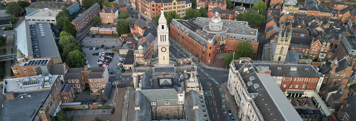 Aerial view of a town with prominent buildings, including a church with a clock tower, surrounded by streets and smaller structures. The area features a mix of modern and historic architecture.