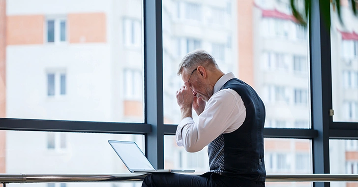 A person in business attire sits at a desk with a laptop, appearing stressed or thoughtful, in front of a large window with a cityscape view.