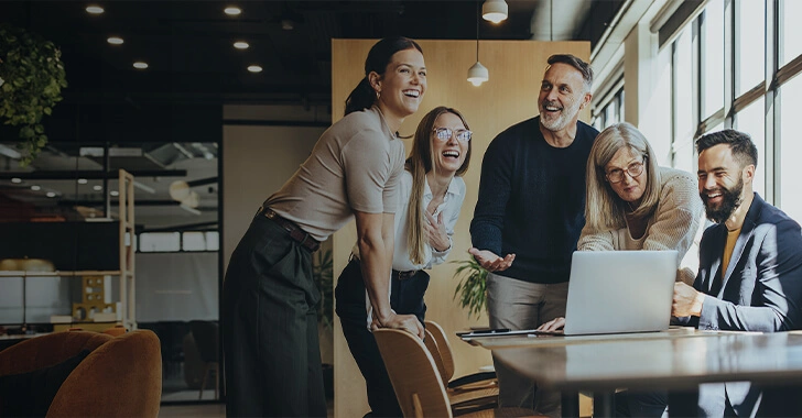 A group of people joyfully collaborating around a laptop in a modern office setting with large windows and indoor plants.