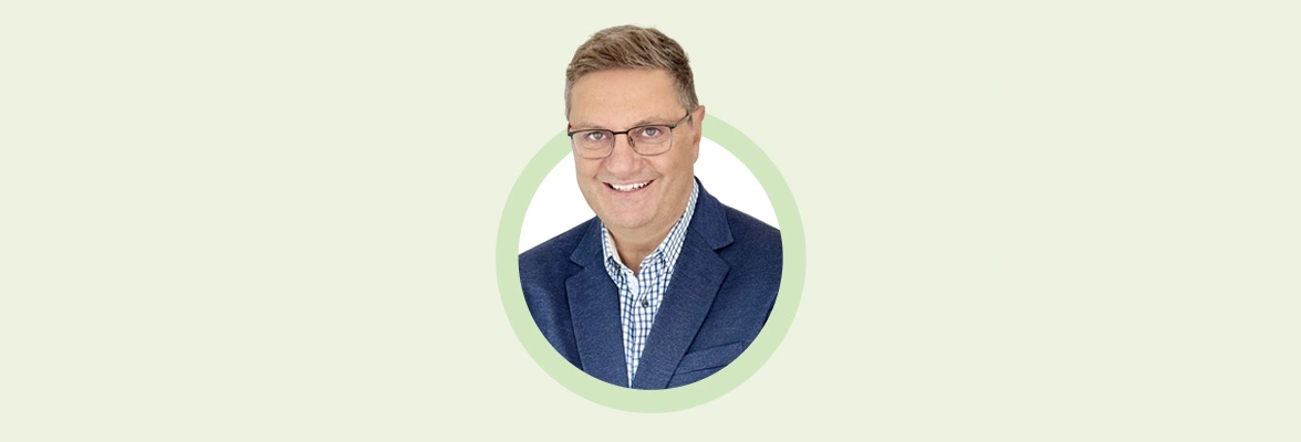 Portrait of Derek Kudsee, smiling and wearing glasses, a blue blazer, and a checkered shirt, against a light background.