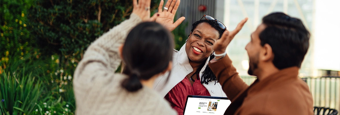 Three people engage in a cheerful conversation outdoors, with one person high-fiving another while the third person holds an open laptop.