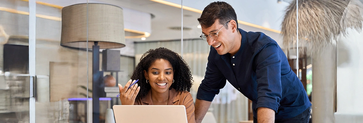Two people are smiling and looking at a laptop in a modern office setting. They are sitting and standing at a table with papers. The environment is bright with a large window and contemporary decor.