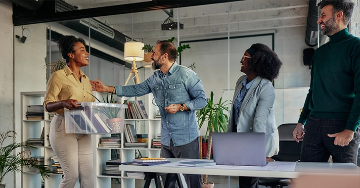 Two professionals shaking hands in an office setting with two other colleagues watching and smiling.