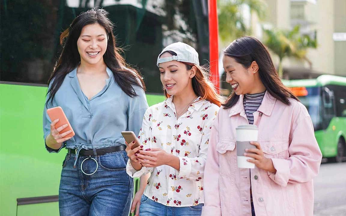 3 girls looking at smartphones and laughing in front of a bus