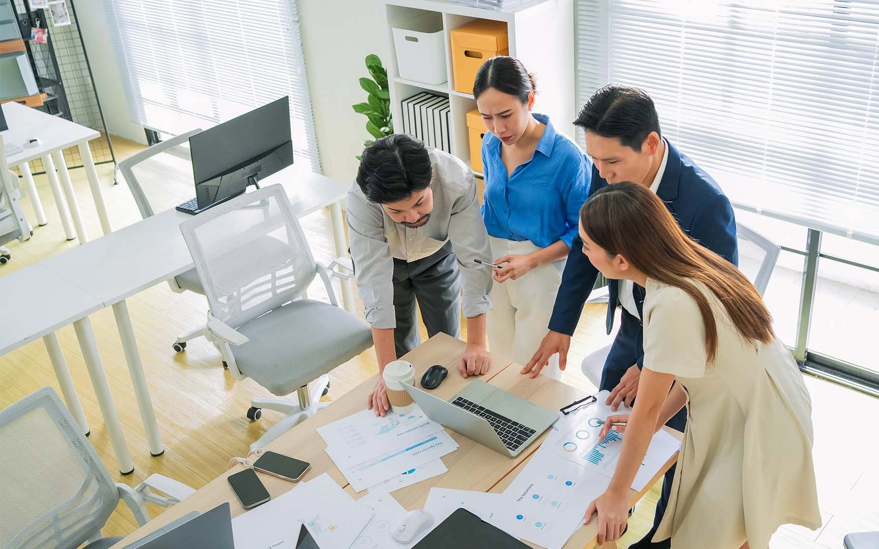 Four professionals gather around a desk in a bright office, reviewing charts and documents alongside a laptop during a collaborative planning and analysis session.