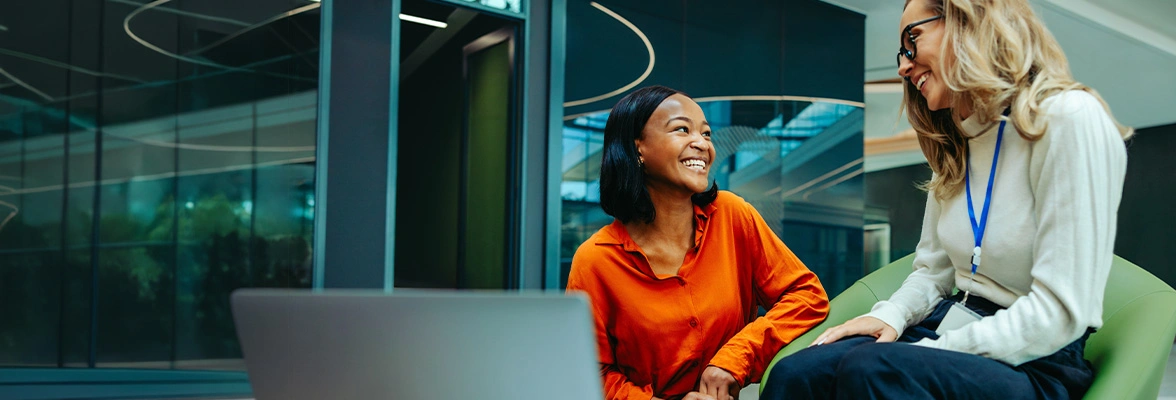 Two people are sitting in a modern office space, engaged in a conversation. Both are smiling. A laptop is open on a table in front of them. The setting has a contemporary design with glass and sleek furniture.