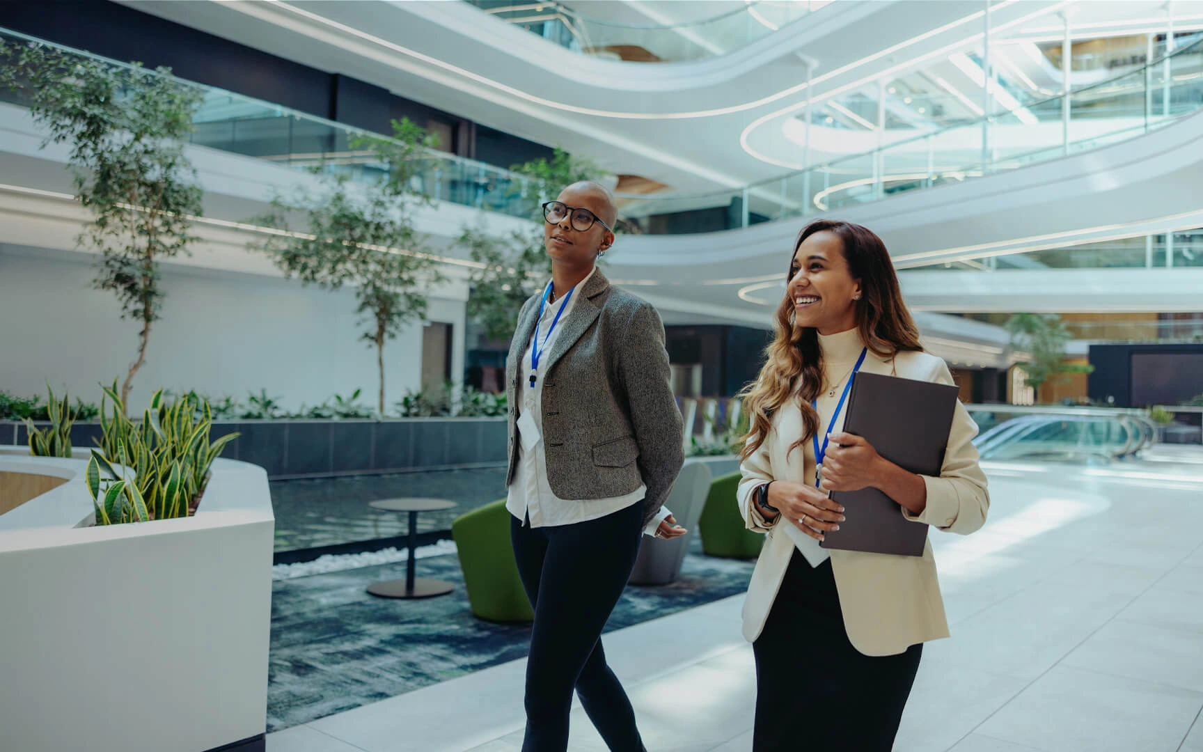 Two professionals wearing visitor badges walk through a bright, modern office atrium with curved architectural features, indoor trees, and collaborative seating areas.