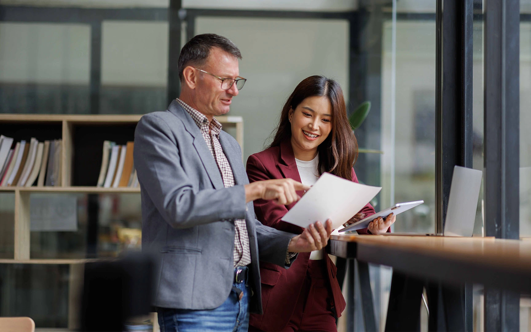 Two professionals standing in a modern office workspace reviewing printed documents together beside a workstation.