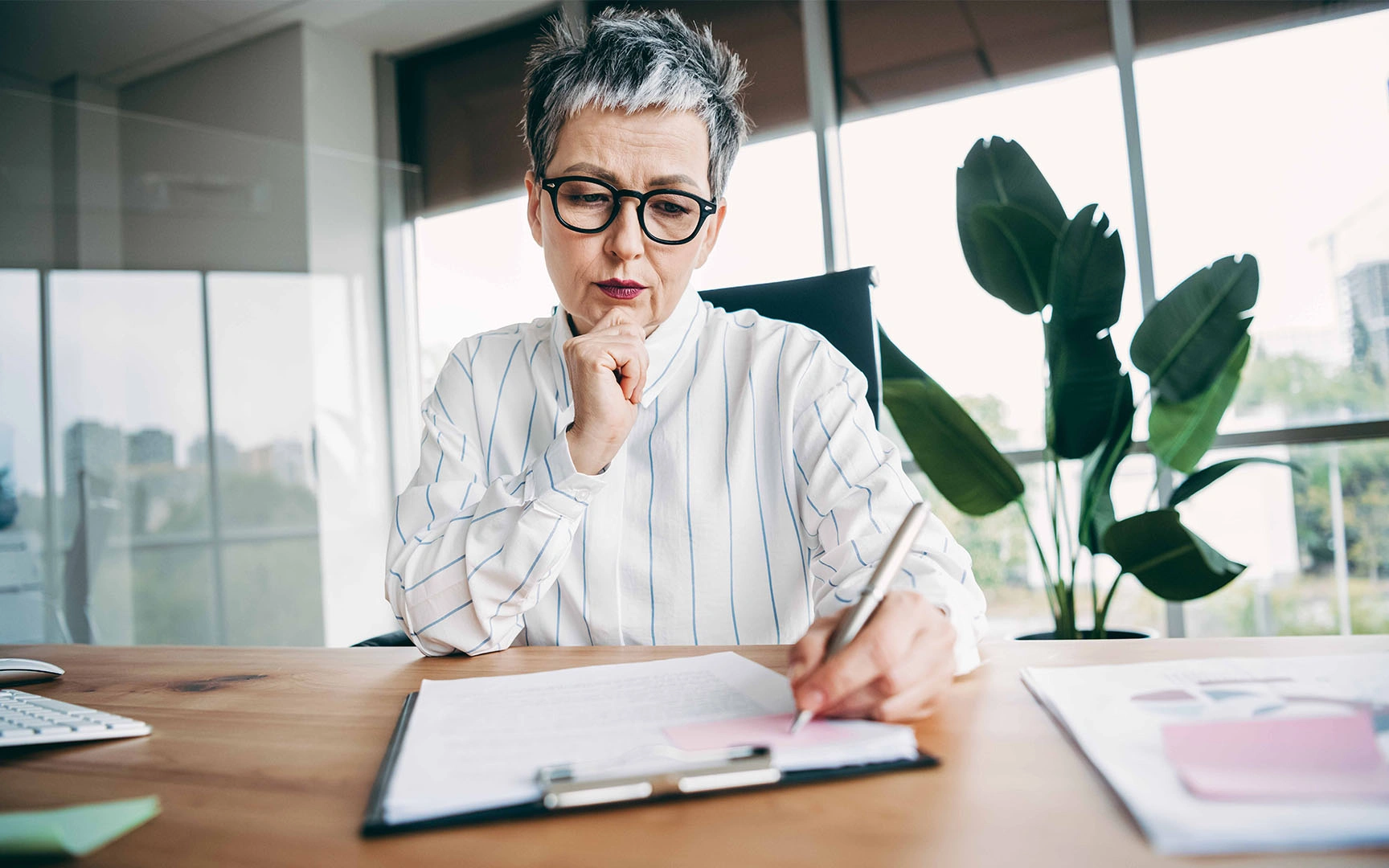 Professional seated at a desk in a modern office, reviewing documents and making notes on a clipboard.