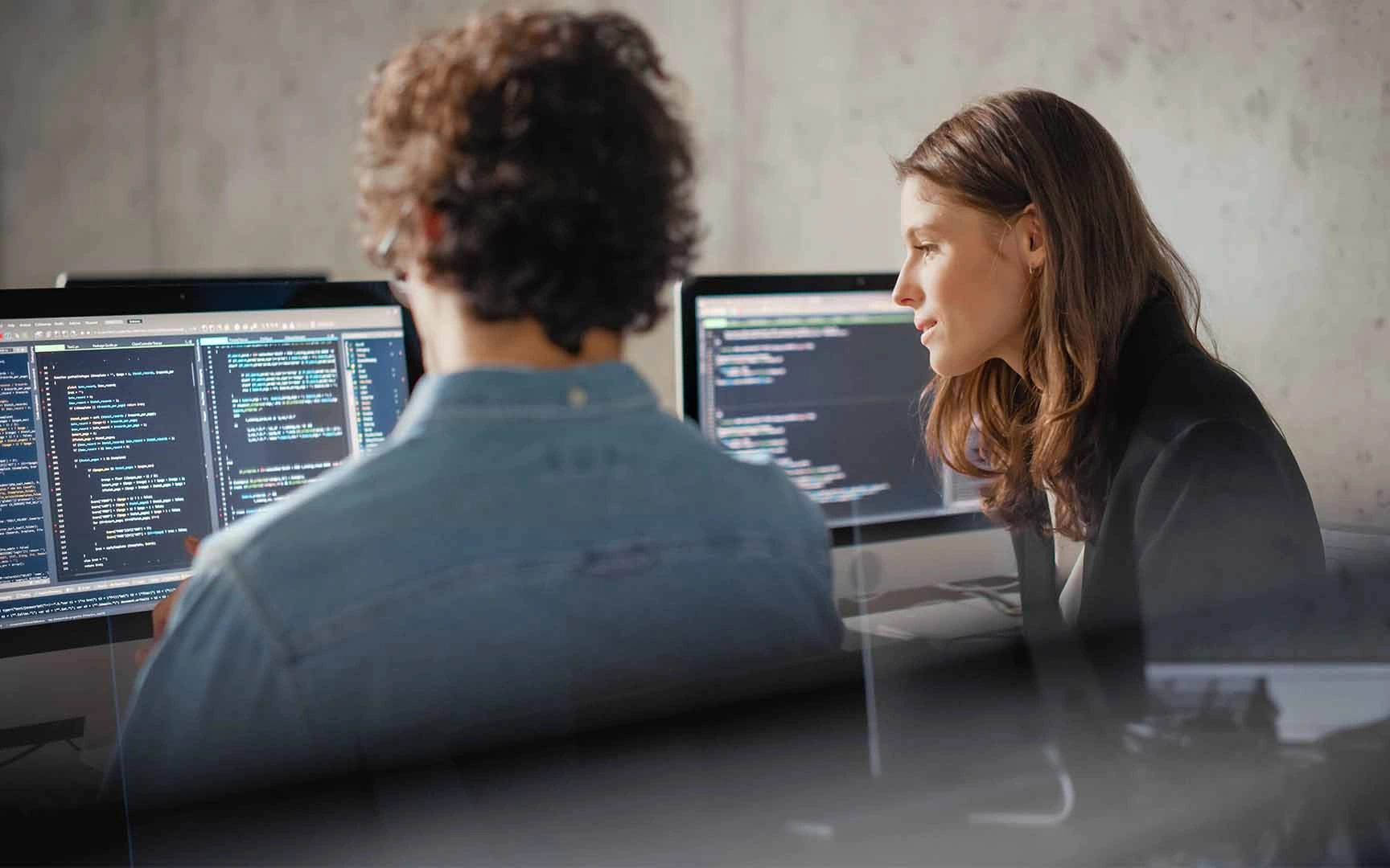 Two professionals collaborating at a workstation, reviewing software code displayed on multiple computer monitors in a modern office environment.