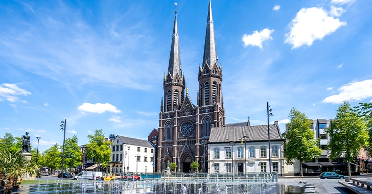 Stadsplein in Tilburg met fontein en de Heuvelse Kerk op de achtergrond, omringd door historische gebouwen en groen onder een blauwe lucht.