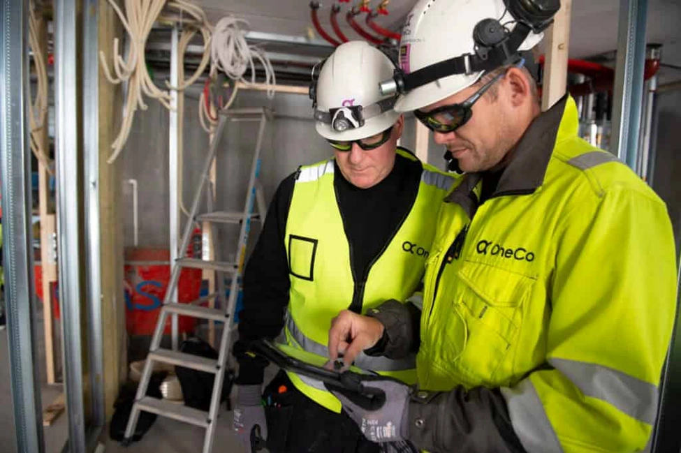 Two construction professionals wearing high-visibility jackets, safety helmets, gloves, and protective eyewear review information on a tablet inside a building site.
