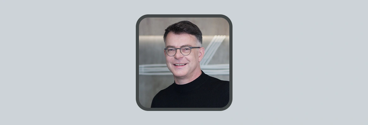 Professional headshot of a person wearing glasses and a dark high-neck top, standing in front of a modern office background with subtle geometric line details.