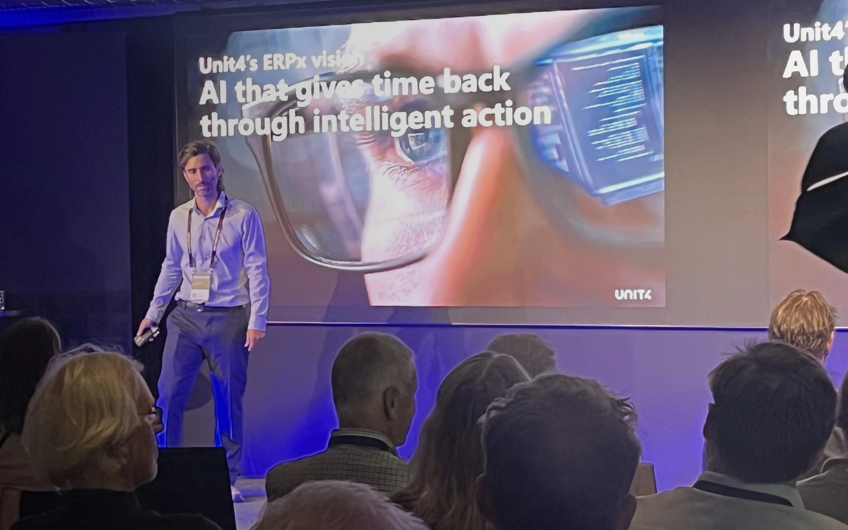 Presenter speaking on stage at a business conference while attendees watch from the audience.
