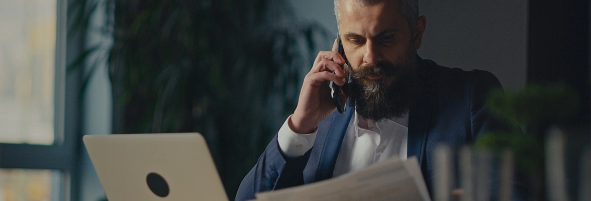 A person with a beard is sitting at a desk, talking on a phone, and holding papers. A laptop is open in front of them. The room is dimly lit.