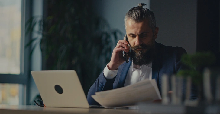 A person with a beard is sitting at a desk, talking on a phone, and holding papers. A laptop is open in front of them. The room is dimly lit.