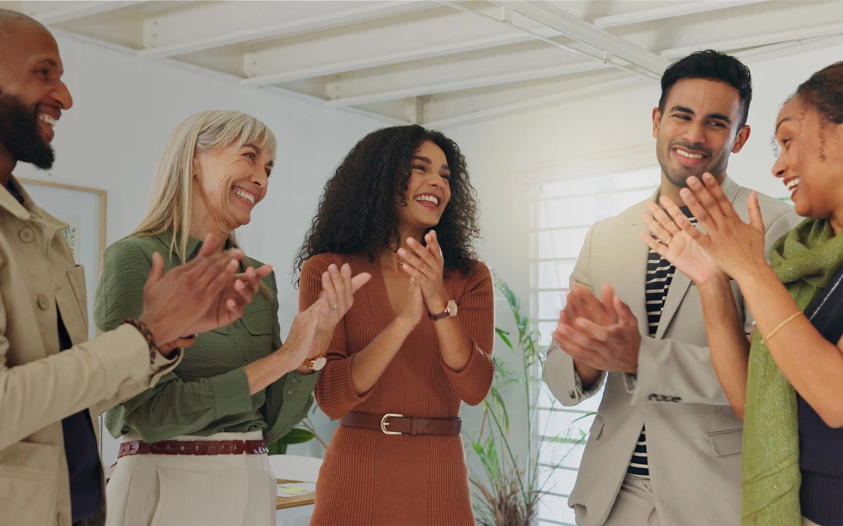A group of five colleagues standing together in a bright office, smiling and applauding during a team gathering.