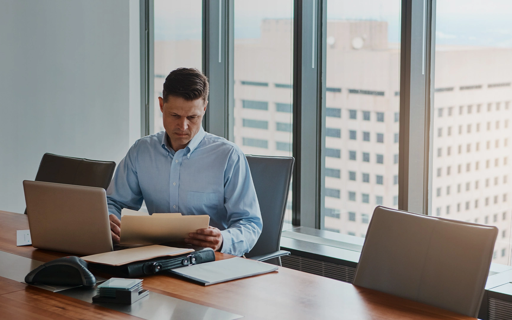 A person in a blue shirt sits at a conference table reviewing documents beside an open laptop, with large office windows and buildings visible in the background.