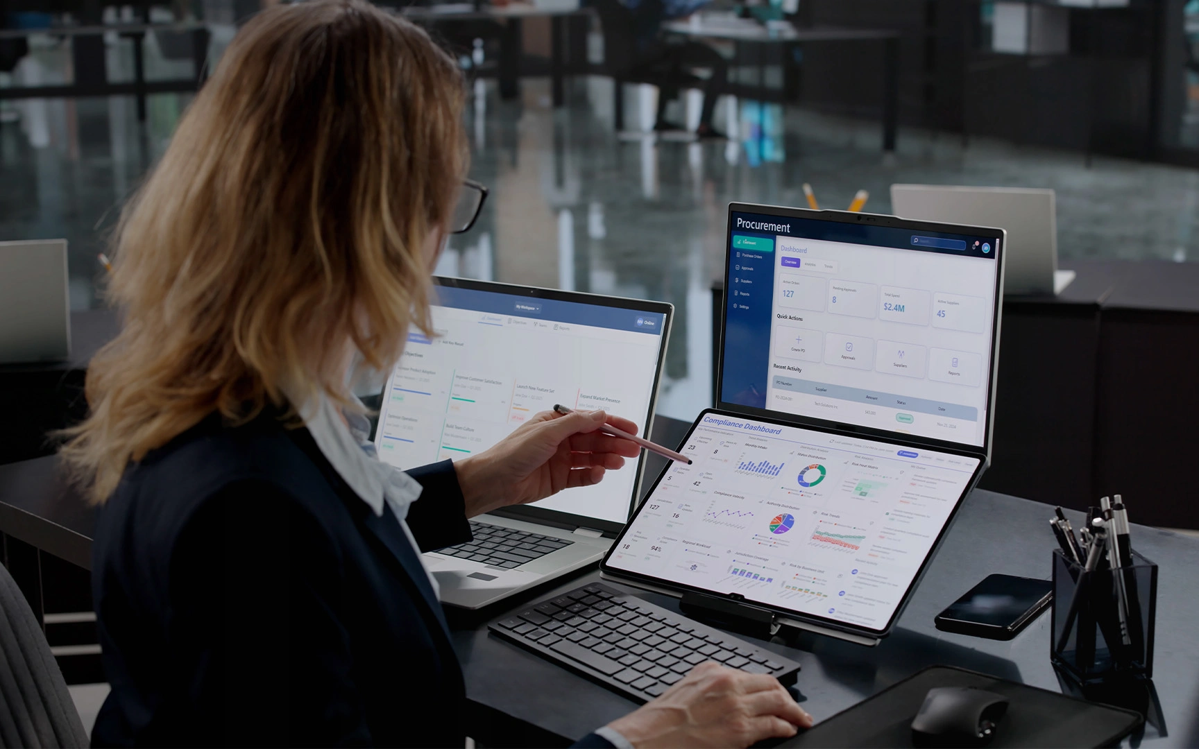 Person reviewing procurement and compliance dashboards across a laptop, monitor, and tablet in a modern office workspace.