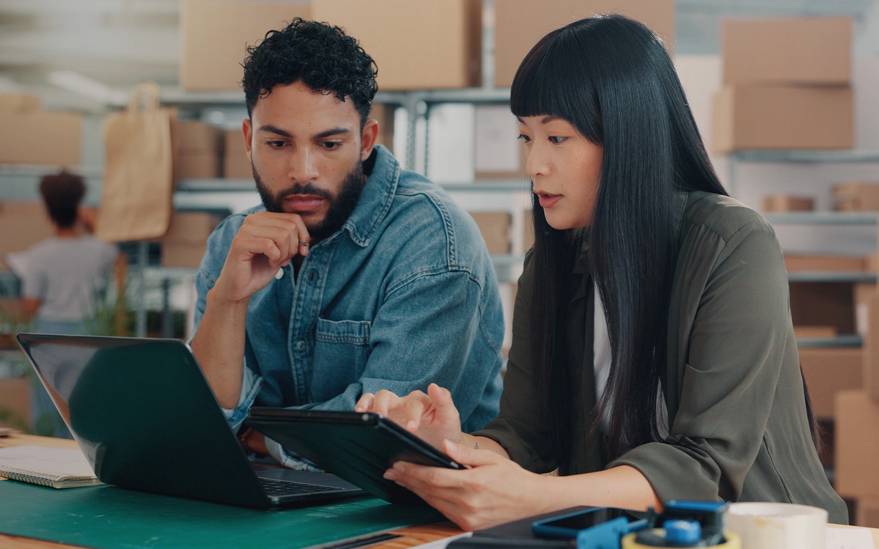 Two colleagues reviewing information on a tablet and laptop together in a modern office workspace.